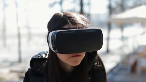Serious-young-dark-haired-woman-in-warm-black-waistcoat-wearing-virtual-reality-glasses-in-early-autumn-park